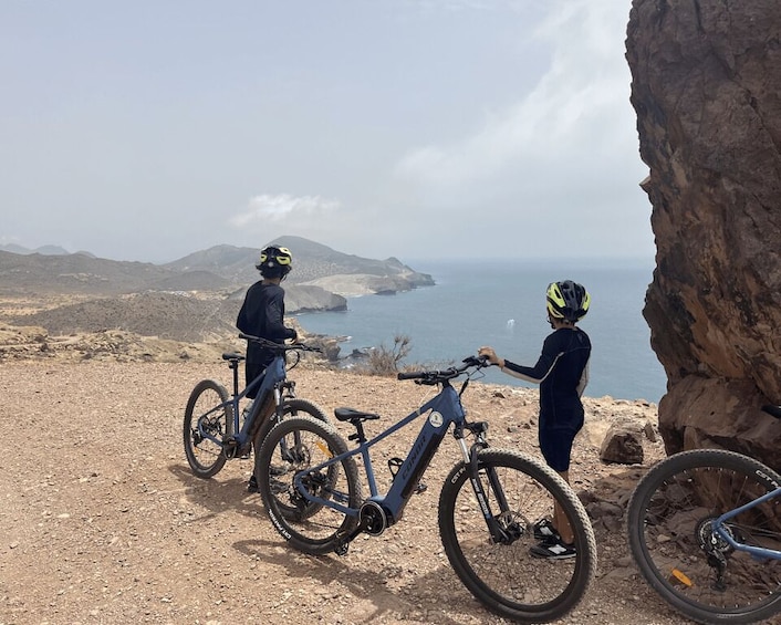 Two cyclists on a rocky terrain with a view of the sea and mountains in the background. 1