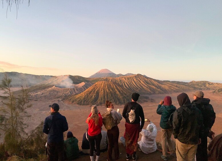 A group of people observing a volcano with smoke rising from its crater. 1