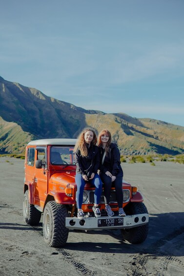 Two people sitting on the front of a red vehicle with a license plate N 1538 SH, parked on a sandy area with mountains in the background. 2