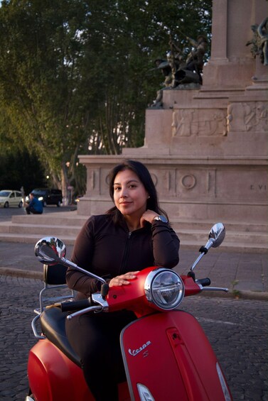 Une personne est assise sur un scooter Vespa rouge devant un monument surmonté d’une statue. 4