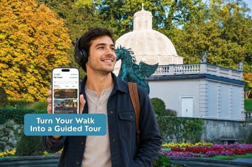 A person holding a smartphone with a tour app, standing in front of a white building with a dome, surrounded by greenery and flowers. 1