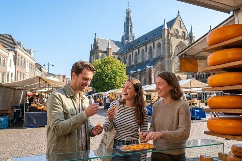 Three people at a market with cheese stands, a church, and cobblestone streets in the background. 1