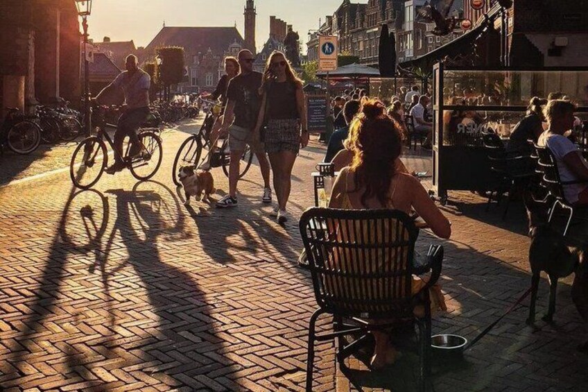 A cobblestone street with people walking and cycling, a dog, and outdoor seating areas. 2