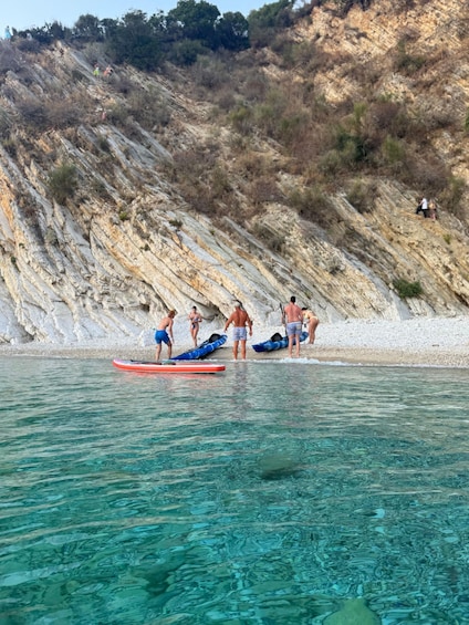 A beach with people preparing to enter the water, a rocky cliff in the background, and clear blue water. 2