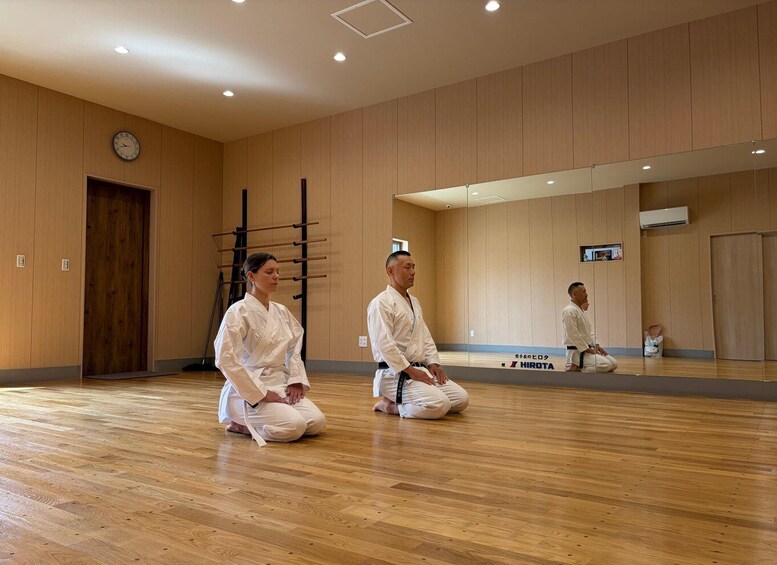 Two individuals in white martial arts uniforms kneeling on a wooden floor in a room with wooden paneling and a large mirror. 1