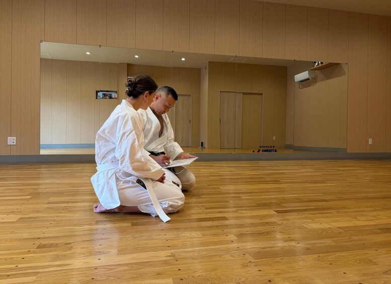 Two people in white martial arts uniforms sitting on the floor of a room with wooden flooring and walls. 3