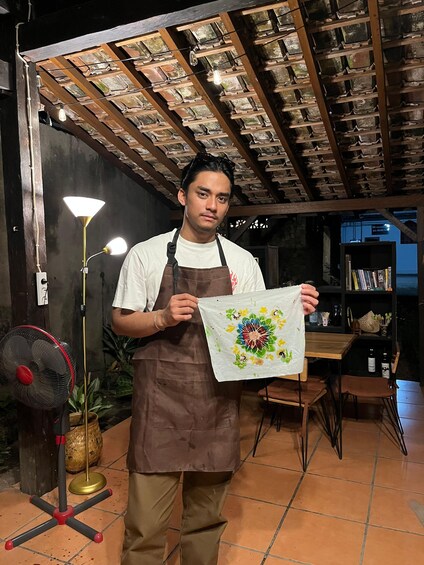 A person holding a colorful paper with a floral design, standing in a room with a tiled floor, a fan, a lamp, and a bookshelf. 5