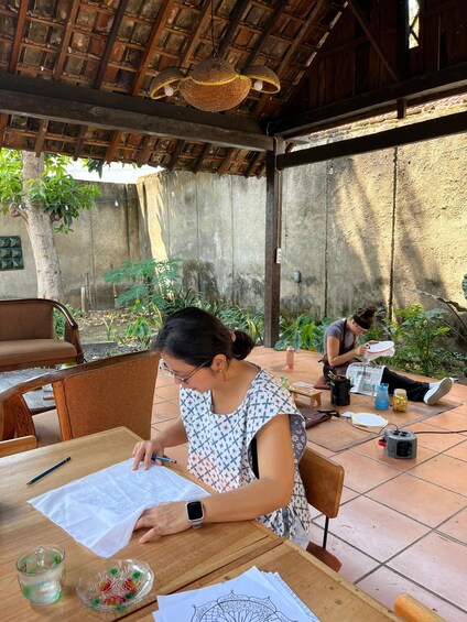 A person sitting at a wooden table outdoors, working on a paper with a pen. 4