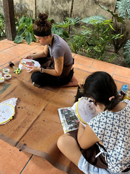 Two individuals engaged in embroidery on a woven mat outdoors, surrounded by greenery. 3