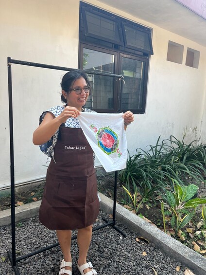 A person holding a white cloth with a colorful floral design, standing in front of a window with a metal frame, wearing a brown apron, and standing on a gravel surface with plants in the background. 2