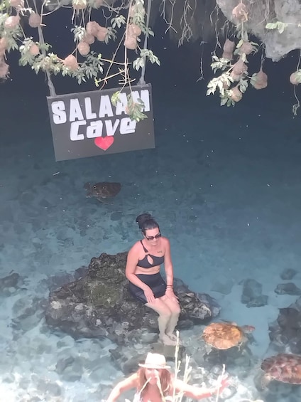 A person sitting on a rock in a clear pool with turtles, a sign that reads 'SALAM COVE', and hanging decorations. 2