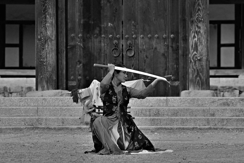 A person in traditional attire holding a sword in front of a wooden building with steps. 4