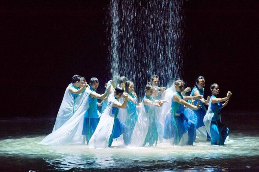A group of dancers in blue and white costumes performing on a stage with a water feature. 1