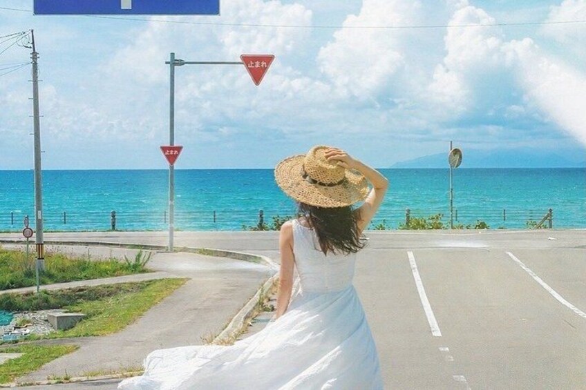 A woman in a white dress and straw hat standing on a road with a view of the ocean. 1