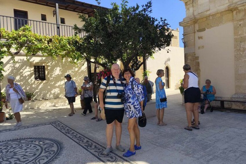 A group of people standing in a courtyard with a patterned rug, a tree, and a building with a balcony. 2