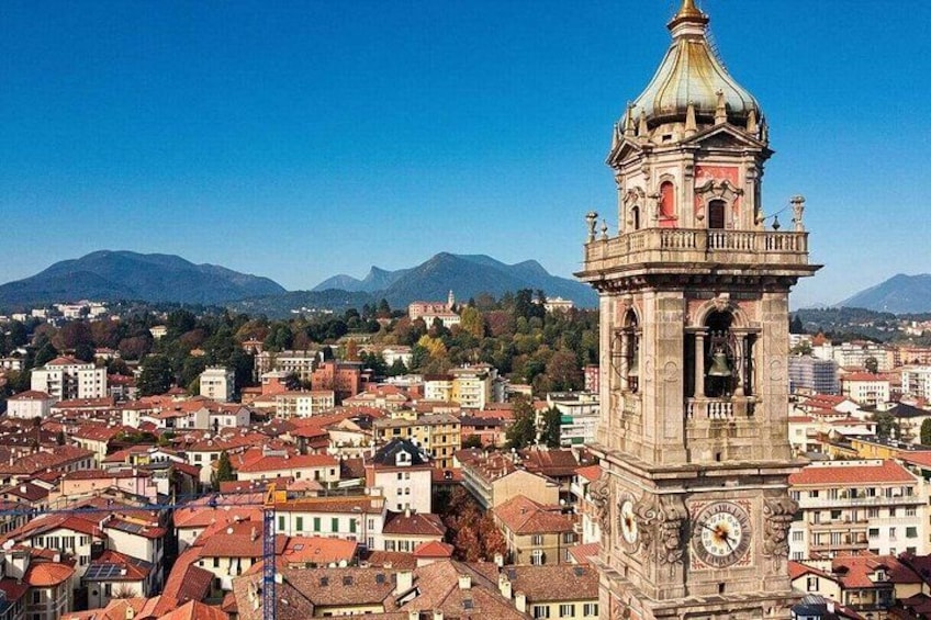 A historic clock tower with intricate architecture, surrounded by a densely built urban area with red-roofed buildings and mountains in the background. 1
