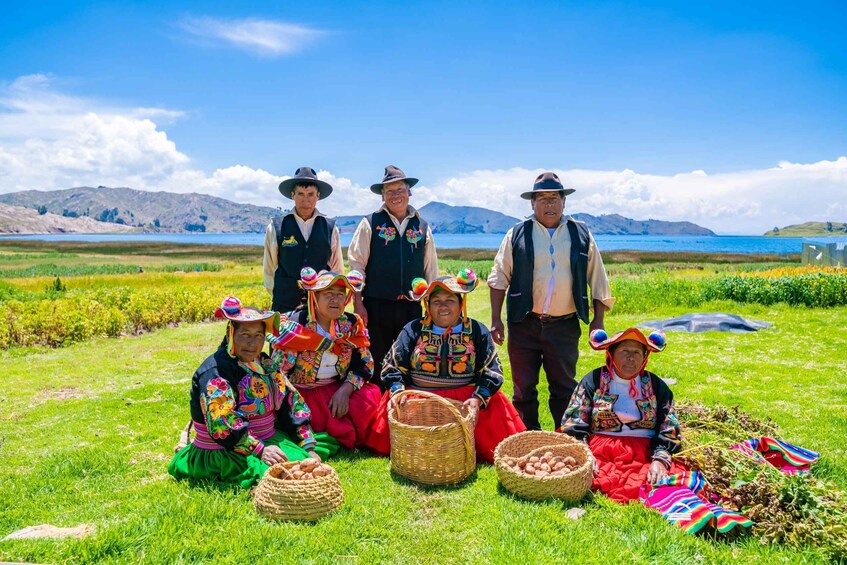 En grupp människor i traditionella dräkter sitter och står på en gräsklädd åker, med en sjö och berg i bakgrunden. 3