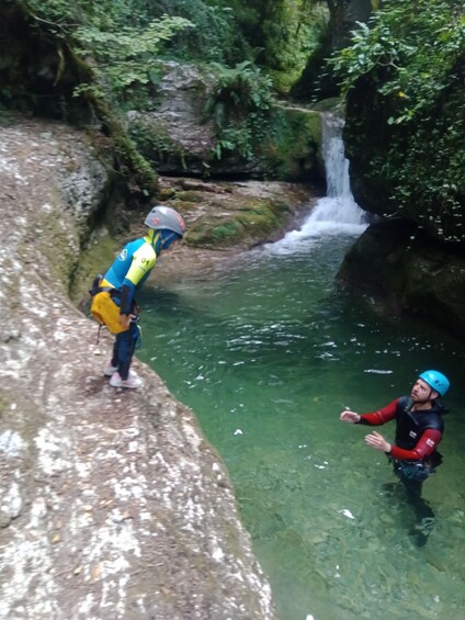 A person in a blue helmet and black wetsuit standing in a river, another person in a yellow helmet and blue jacket on a rock, and a waterfall in the background. 2