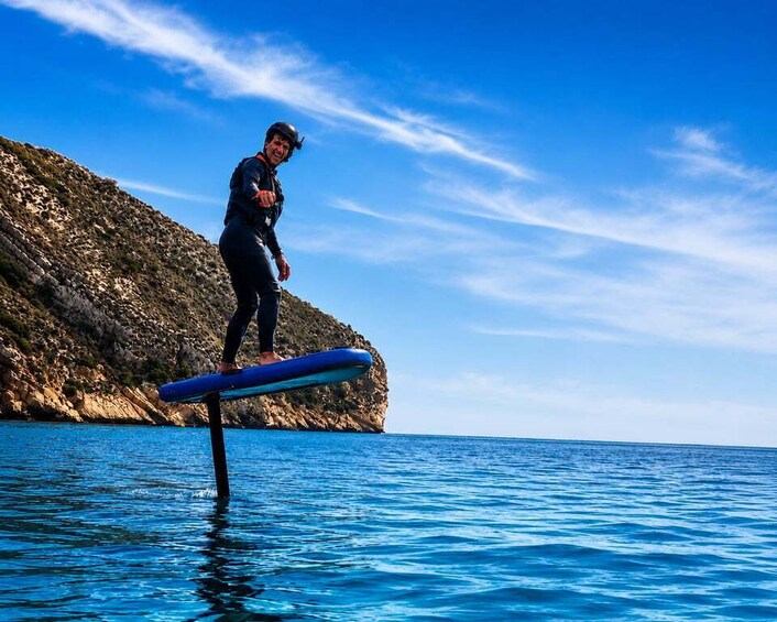 Une personne se tient debout sur une planche de paddle bleu dans l’eau, avec une falaise rocheuse en arrière-plan. 1