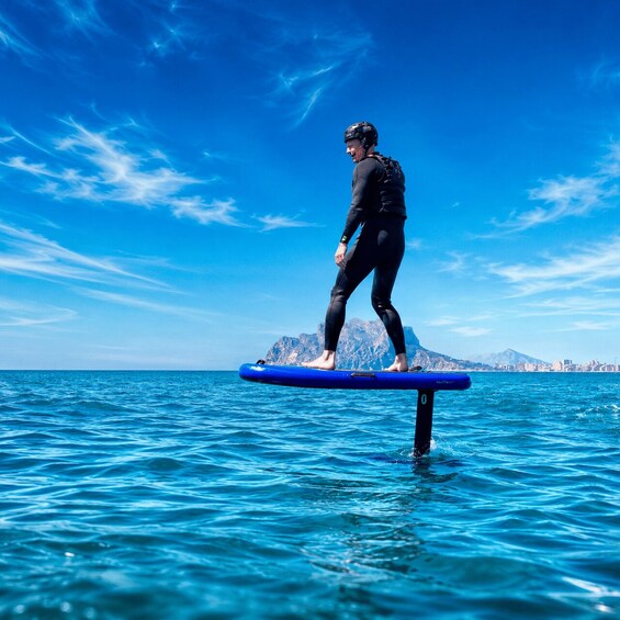 Une personne se tient debout sur une planche de paddle bleu dans l’océan, avec un paysage montagneux en arrière-plan. 5