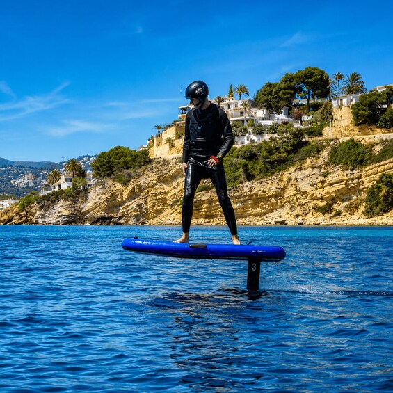 Une personne se tient debout sur une planche de paddle bleu, sur une étendue d’eau, avec un paysage côtier en arrière-plan. 4