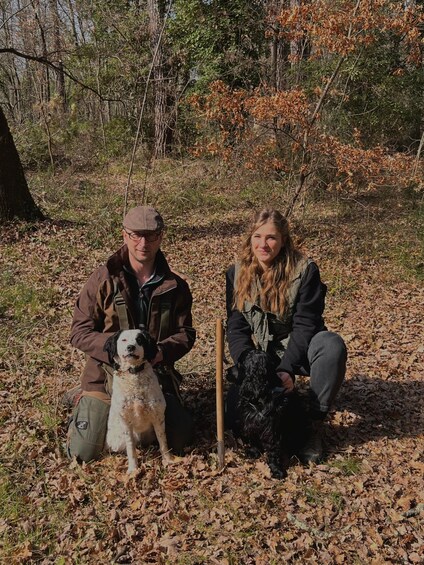Two people sitting on the ground in a wooded area with fallen leaves. 1