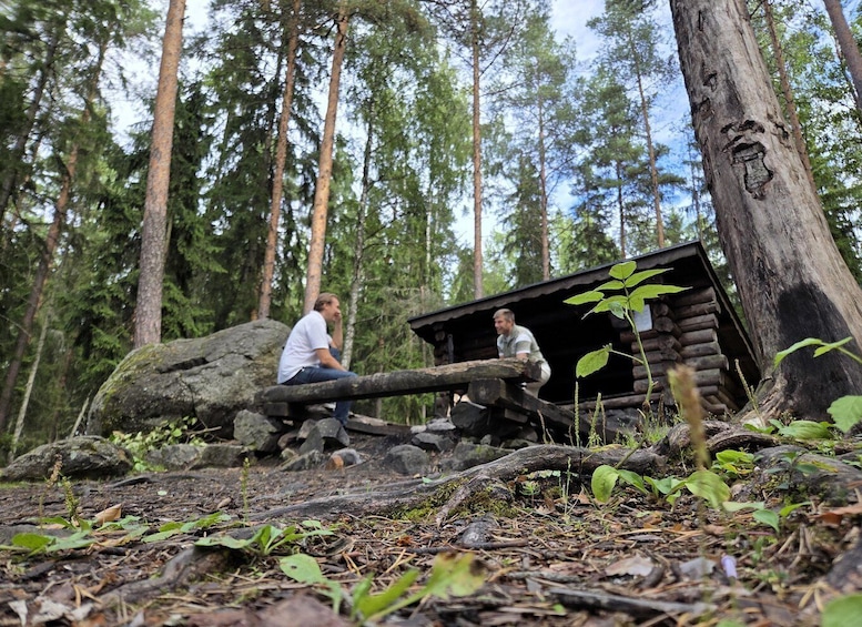 Two people sitting on a log in a forest with a small cabin in the background. 3