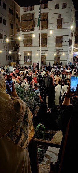 A crowded outdoor gathering with people, some holding cameras, in front of a building with traditional architecture and a flag. 1