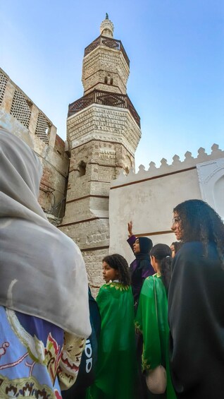 A group of people near a historic tower with intricate stonework and a clear blue sky. 4