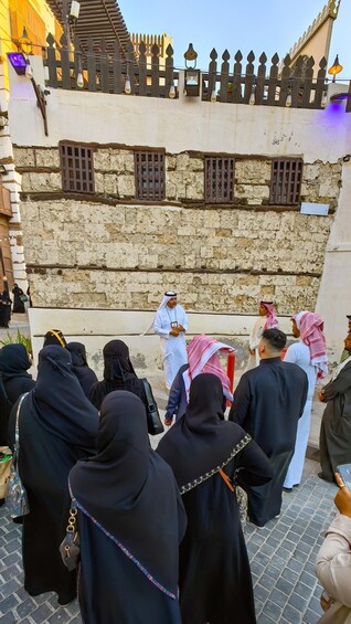 A group of people in traditional attire gathered around a stone wall with barred windows, under a wooden fence with decorative elements. 3