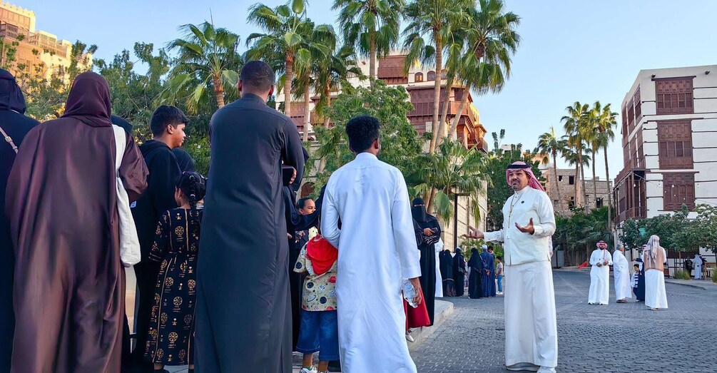 A group of people in traditional attire standing in an outdoor setting with palm trees and buildings in the background. 2