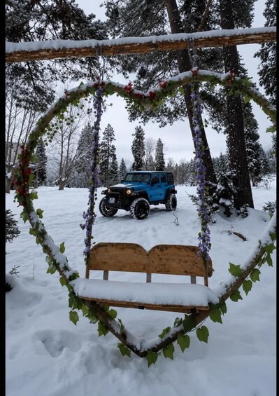 A snow-covered bench surrounded by a heart-shaped wreath, with a blue off-road vehicle parked in the background. 2