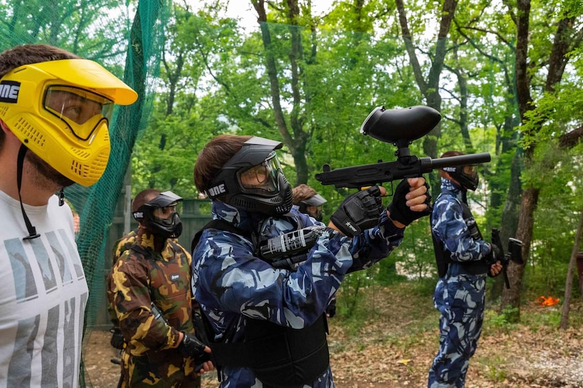 People in camouflage paintball gear, aiming and shooting at targets in an outdoor wooded area. 4
