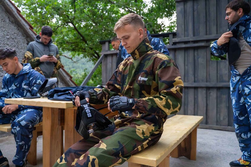 A person in military camouflage attire sitting on a bench, holding a black bag. 2