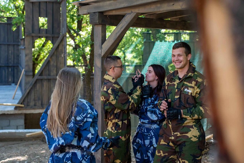 A group of people in military uniforms standing under a wooden structure with a metal mesh roof, surrounded by greenery. 5