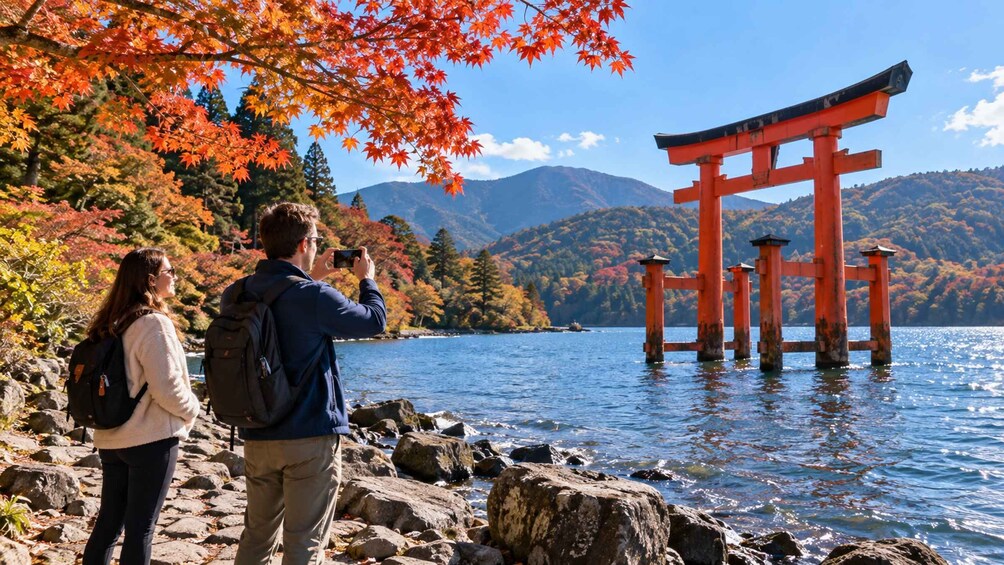 Two people taking a photo of a red torii gate by a lake with autumn foliage. 3