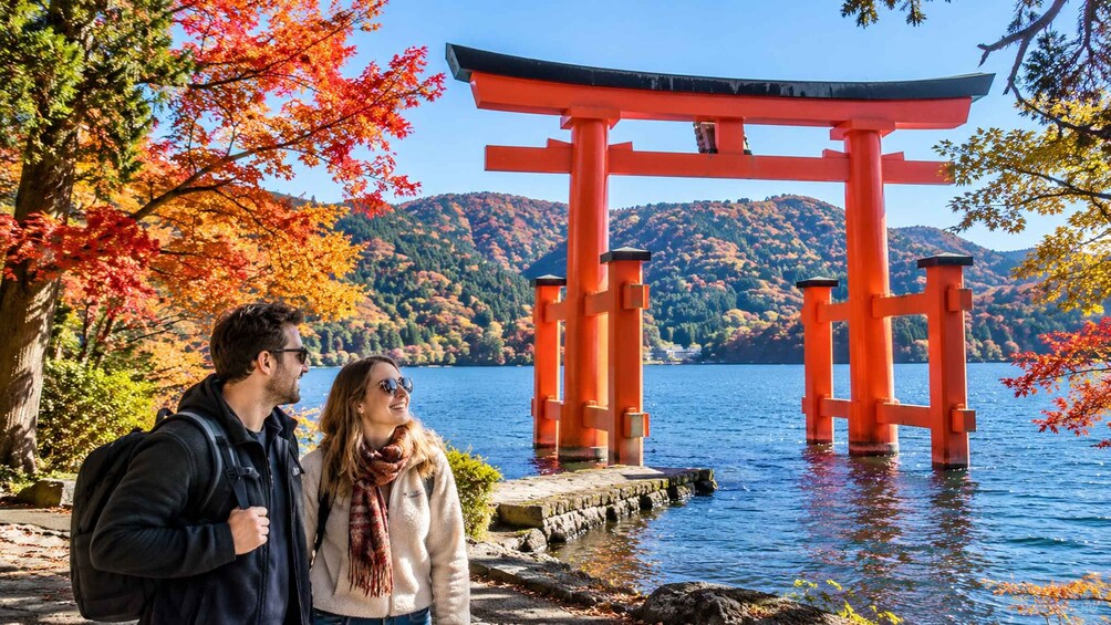 A couple standing by a red torii gate in a scenic autumn setting with a lake and mountains in the background. 1