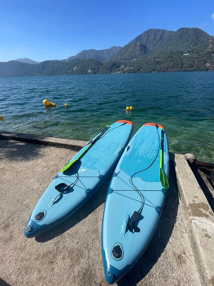 Two blue kayaks with green and orange accents on a concrete dock by a lake with mountains in the background. 2