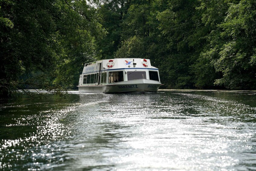 A white boat with 'WAKEWITTZ' written on the side, moving on a calm river surrounded by dense green foliage. 4