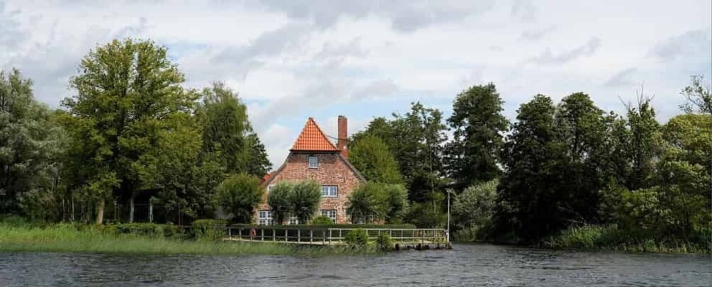 A brick house with a red roof by a river, surrounded by trees and a wooden dock. 3