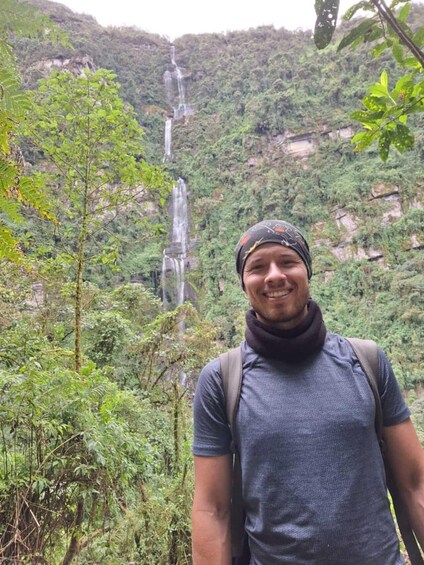 A person standing in a forested area with a waterfall in the background. 2