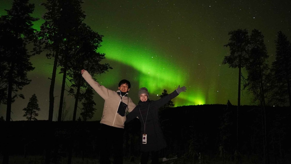 Two people standing with arms raised, facing a night sky with green aurora borealis. 3