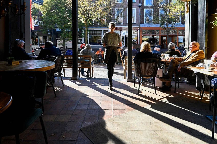 An outdoor cafe with people seated at tables, a woman walking through the space, and buildings in the background. 3