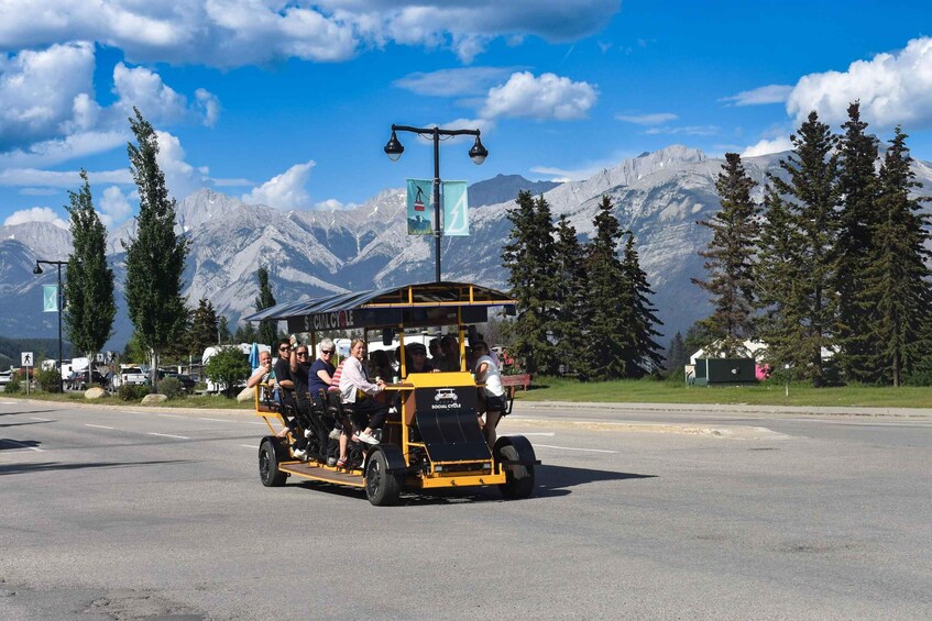 A yellow and black social cycle vehicle with passengers, parked on a road with a mountainous backdrop. 1