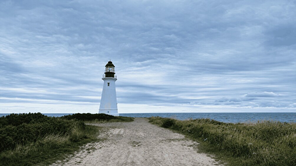 A white lighthouse on a gravel path with grass on either side, leading to the sea under a cloudy sky. 2