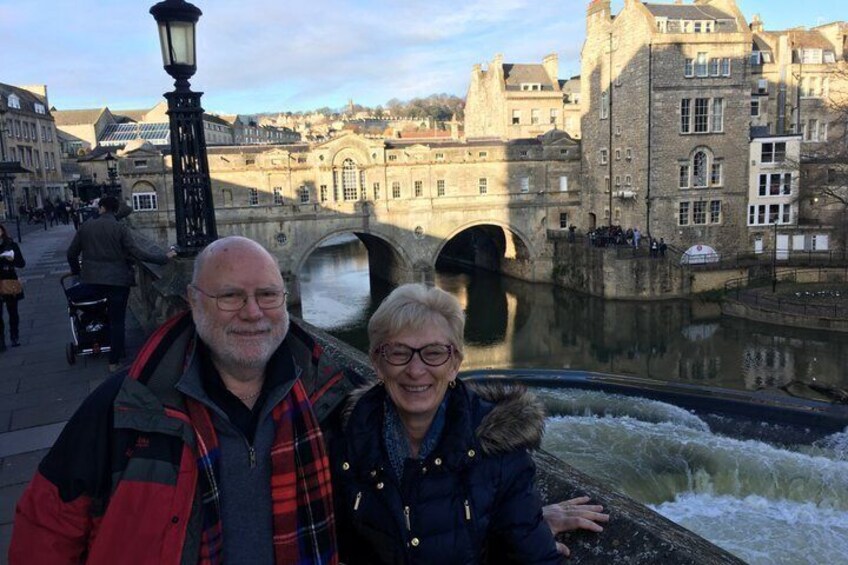 A couple standing by a river with a bridge in the background. 1