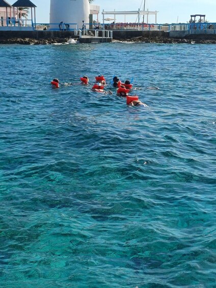 People swimming in a clear blue sea with a pier and buildings in the background. 2