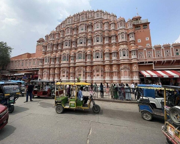 A large pink palace with intricate architecture, surrounded by a busy street with auto-rickshaws and pedestrians. 3