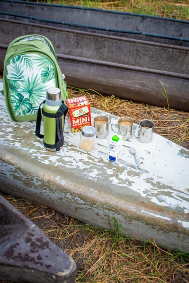 A picnic setup with a green bag, a box of mini rusks, a thermos, a jar of sugar, and several metal cups on a weathered wooden surface. 5