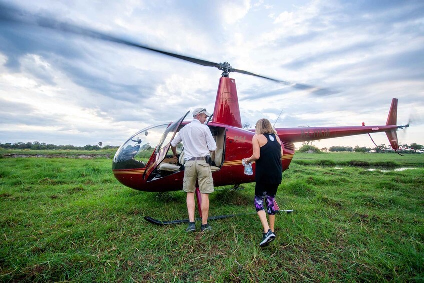 A red helicopter with a person in a white shirt and khaki shorts standing next to it, and another person in a black tank top and purple pants holding a water bottle. 4
